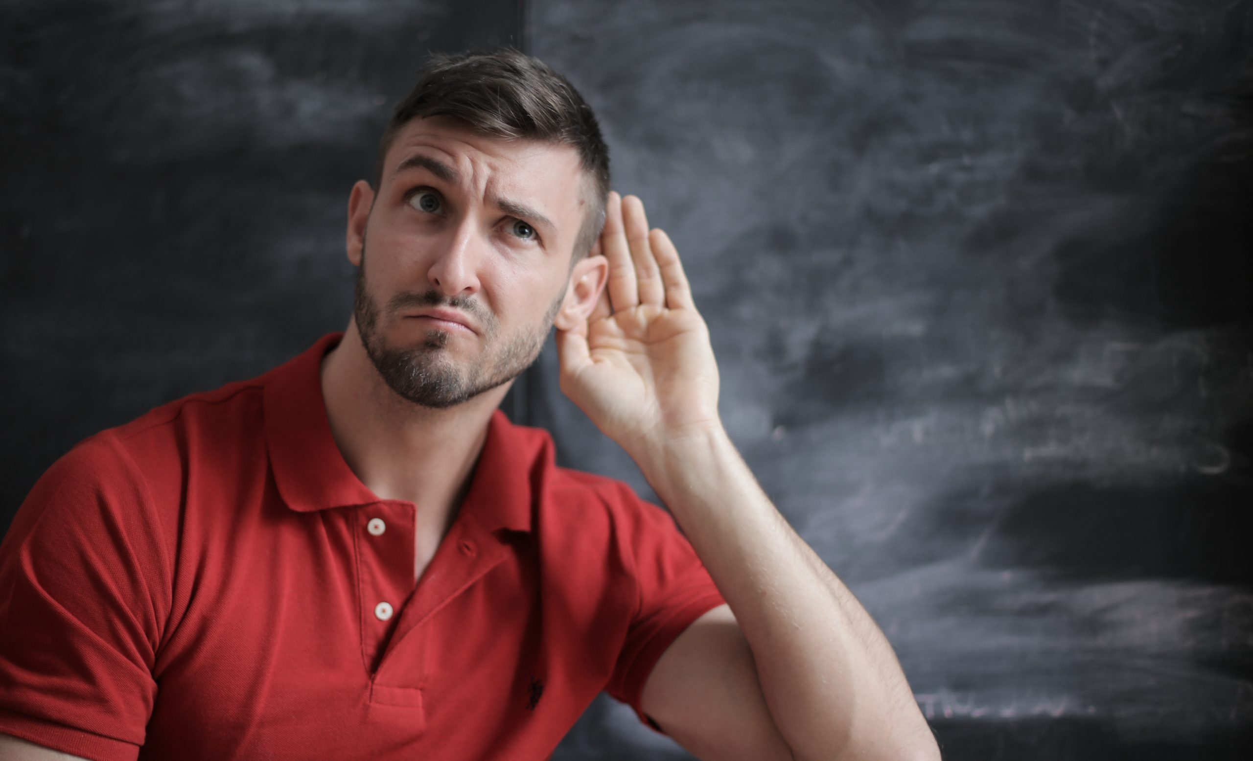 Man in a red shirt holding his hand up to his ear as if he is trying to hear better.