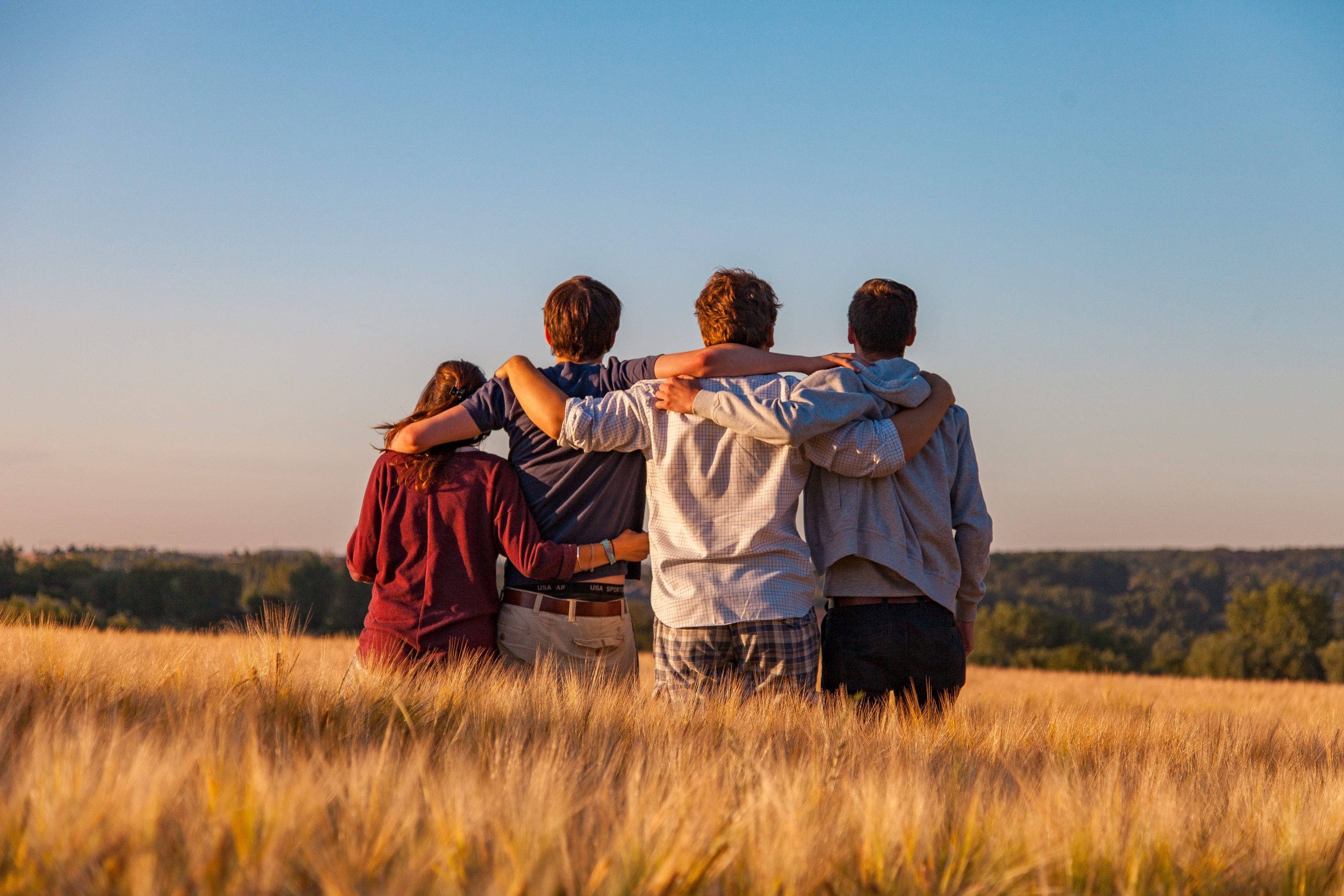 Photograph from behind of 4 people with their arms around each other, standing in a field.