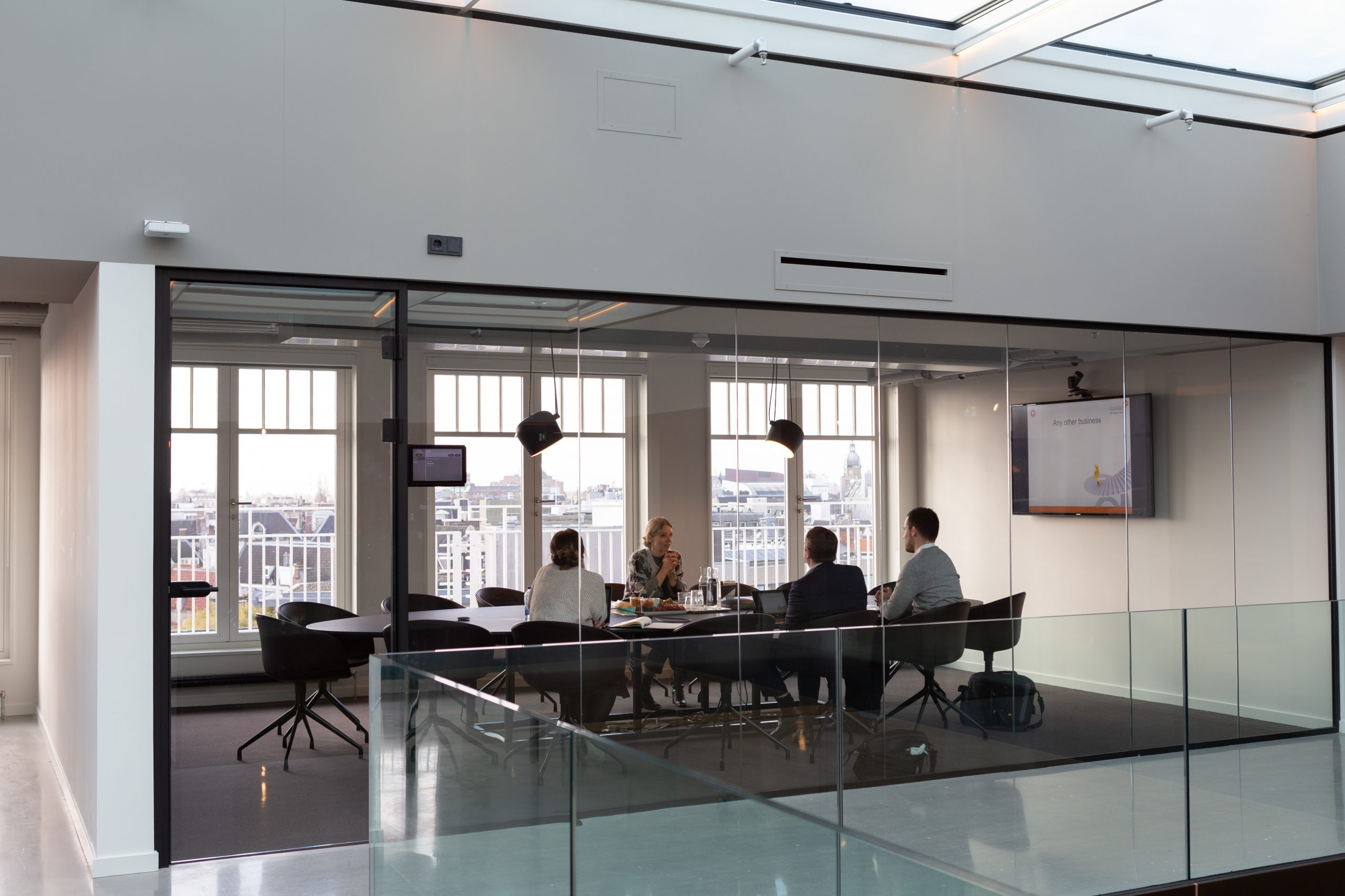 Photograph of a conference room with 4 people sitting at a table. There's a powerpoint on the tv in the background.