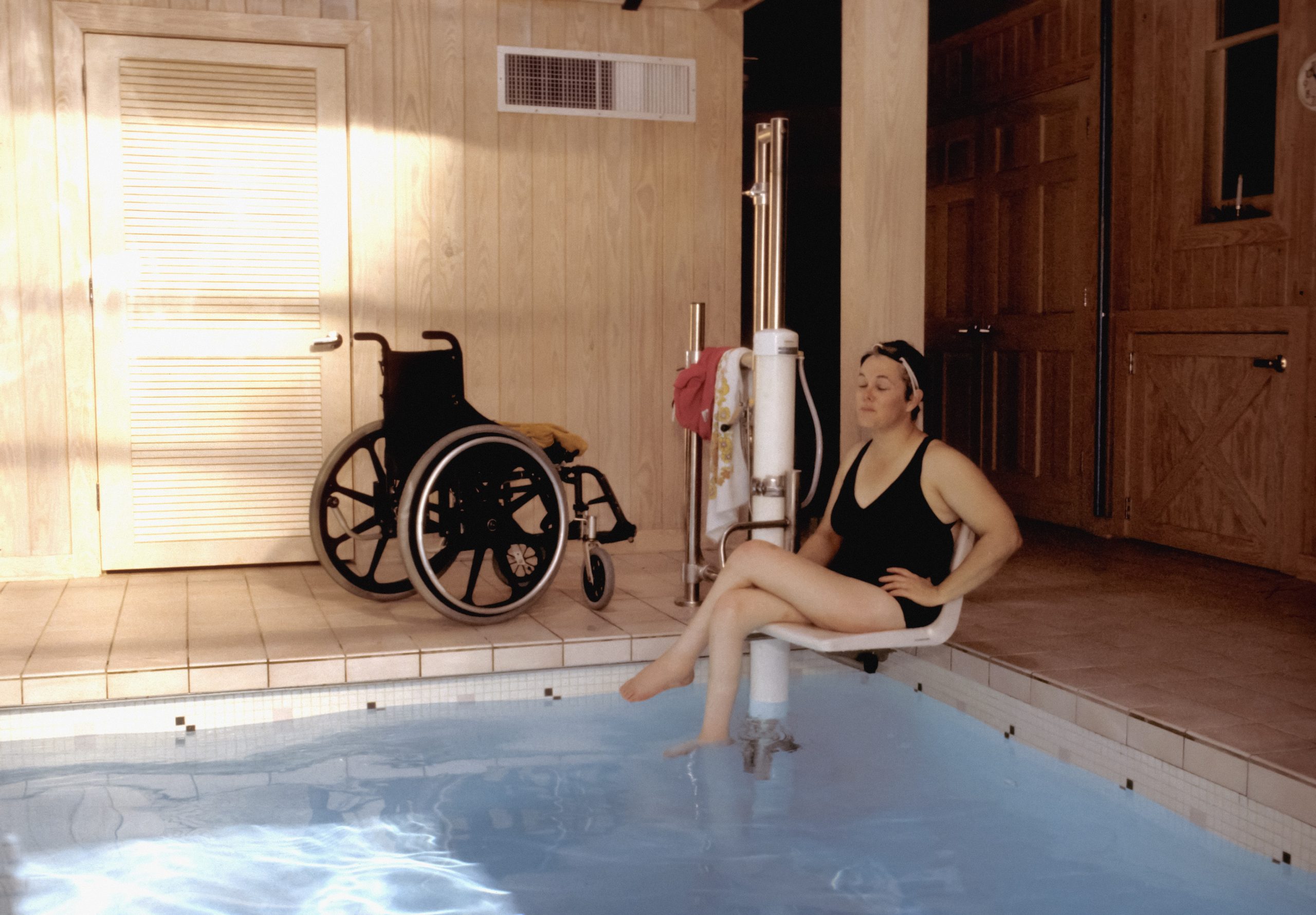 Photograph of a woman using a wheelchair-to-water pool lift to enter a pool. Her wheelchair is in the background.