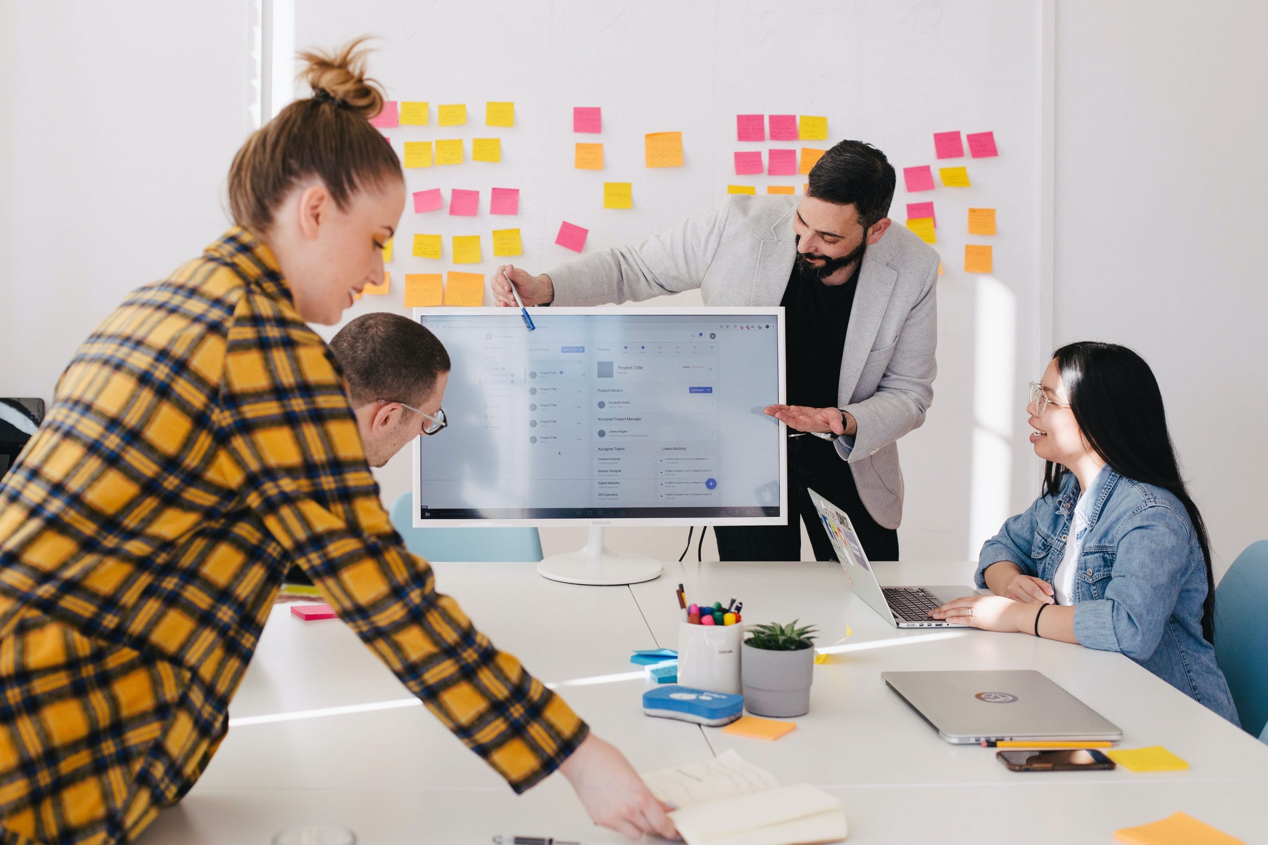 4 people sit at a table, point at a computer screen, and are talking. There's sticky notes on a whiteboard in the background.