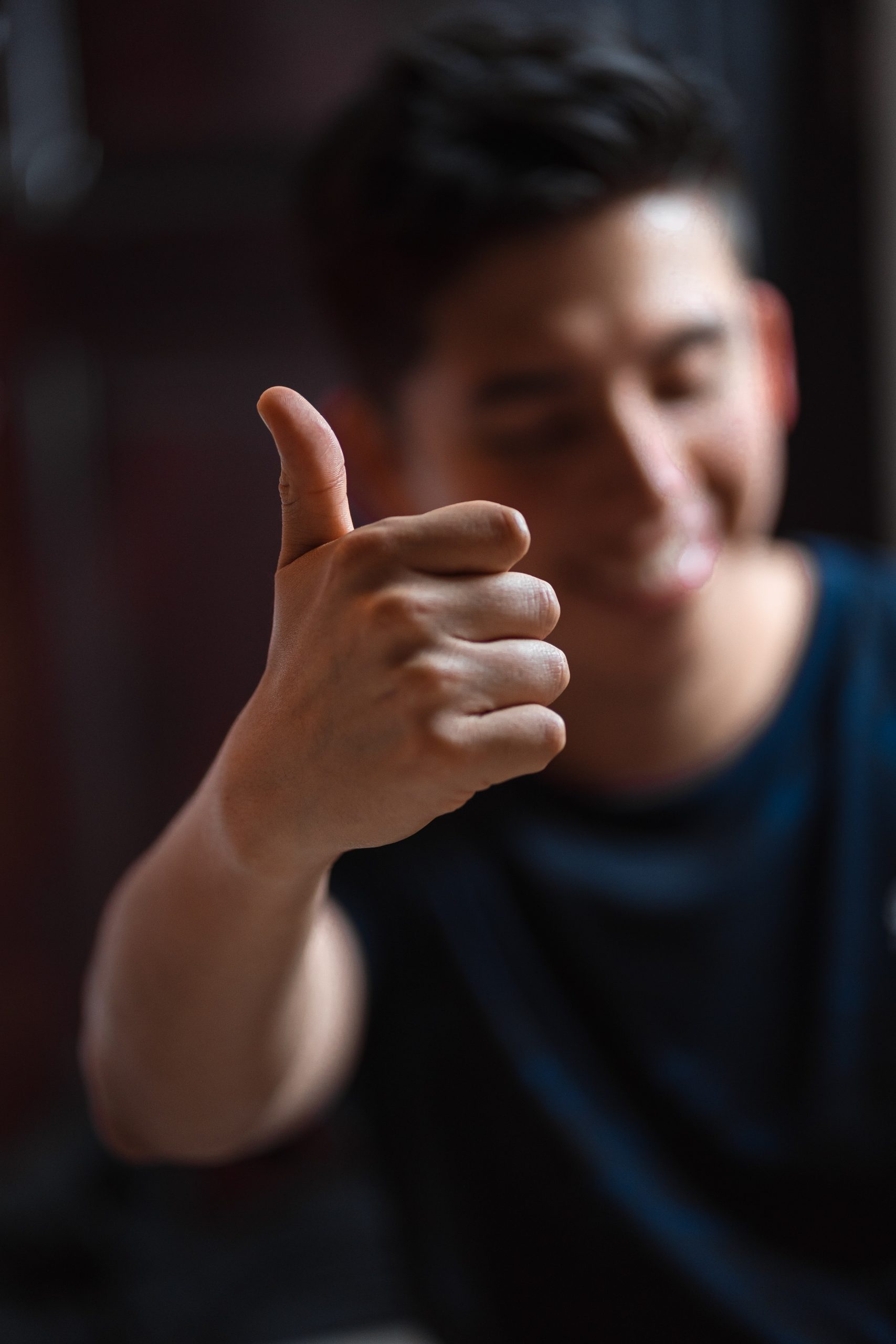 Photograph of a man holding a thumbs up gesture. Only his hand is in focus.