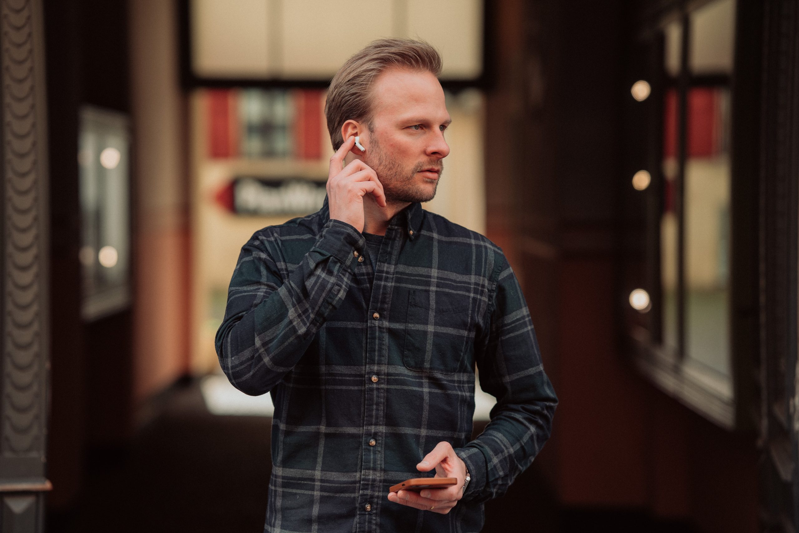 Man in a plaid shirt with a phone in one hand. The other hand is adjusting an Airpod in his ear.