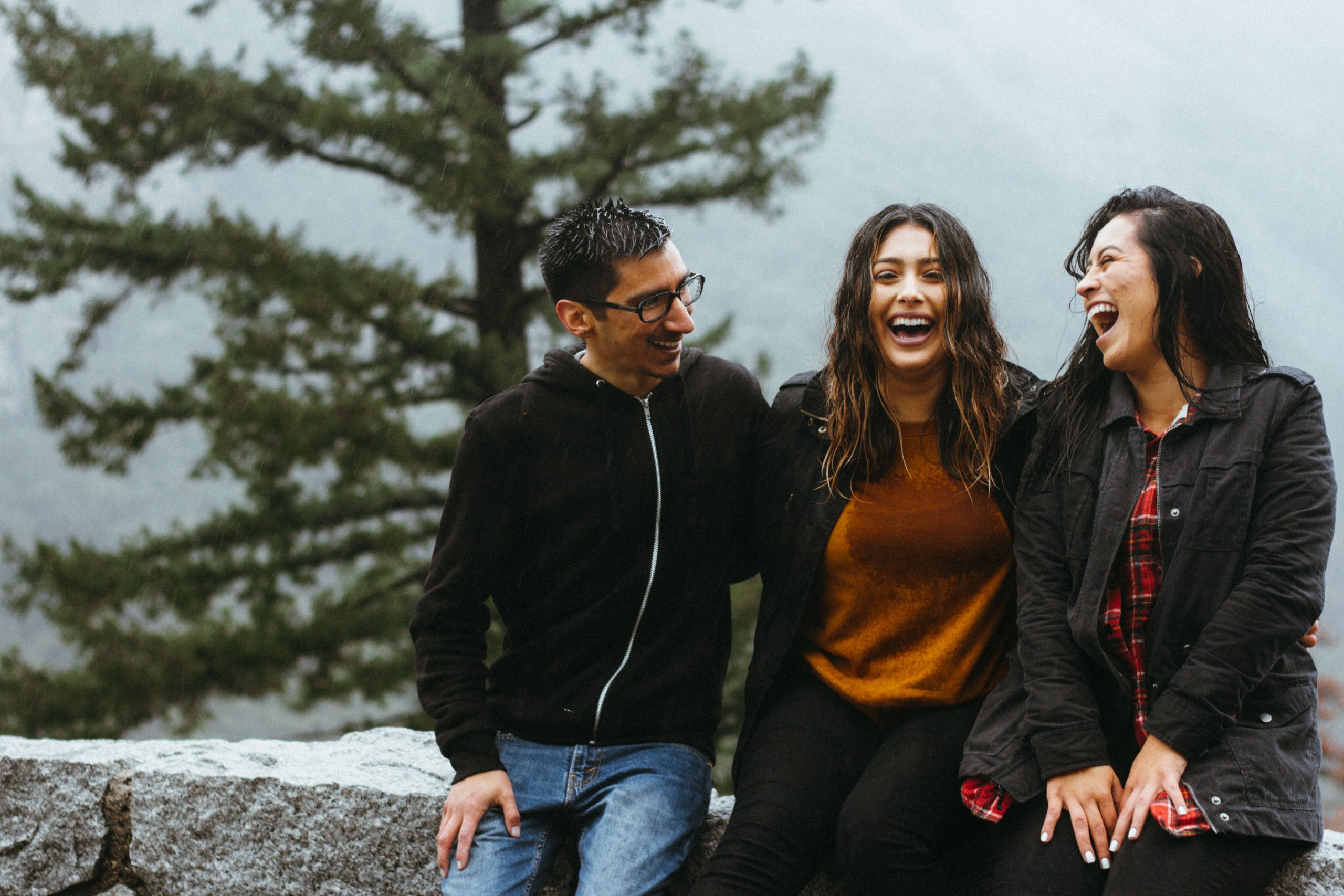 Three people outside sitting on a rock ledge laughing at eachother