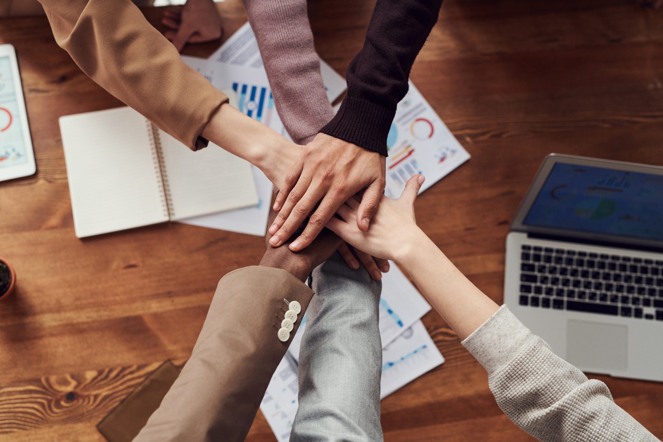 Photo of 6 different hands on top of each other in a circle. Below the hands are papers and laptops on a table.