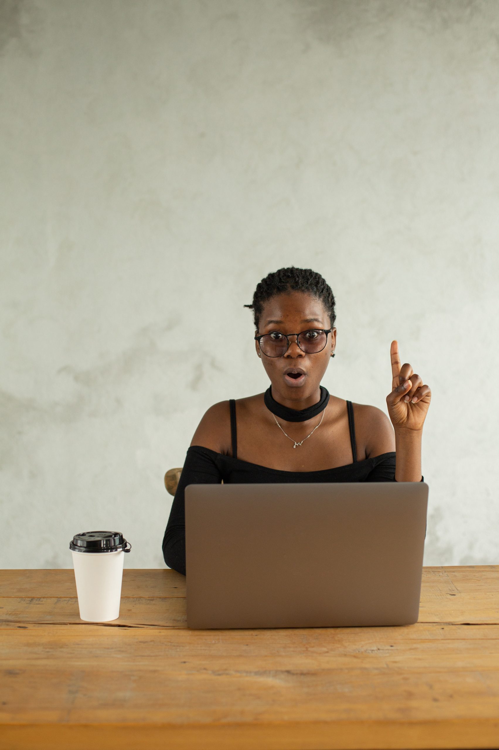 Photo of a woman sitting at her laptop drinking coffee. Her finger is pointed up, eyebrows raised, mouth in an O shape, eyes widened, as if she has a good idea.