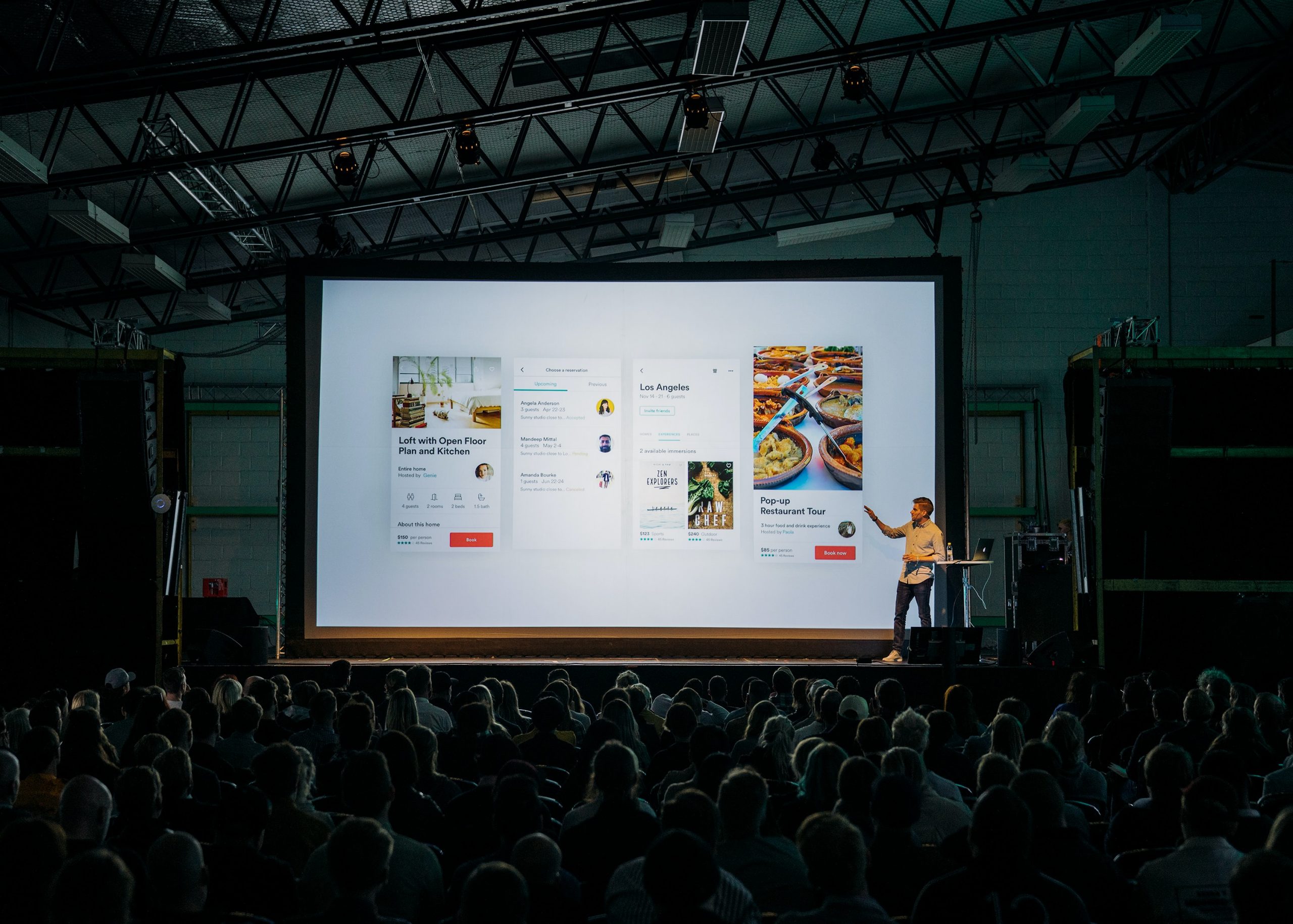 Man on stage pointing to a screen that displays images, descriptions, and fun layouts. There's a large crowd in the audience.