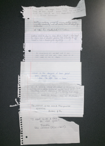 On a black surface rests a cento poem consisting of several ripped pieces of notebook paper, each with different lines of poetry written in different hands, laid out to create "one" long poem.